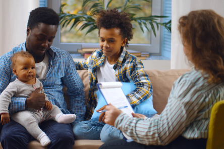 Social worker consulting smiling young single father with kids at home. Case worker woman visiting african man with schoolboy son and baby daughter providing consultation
